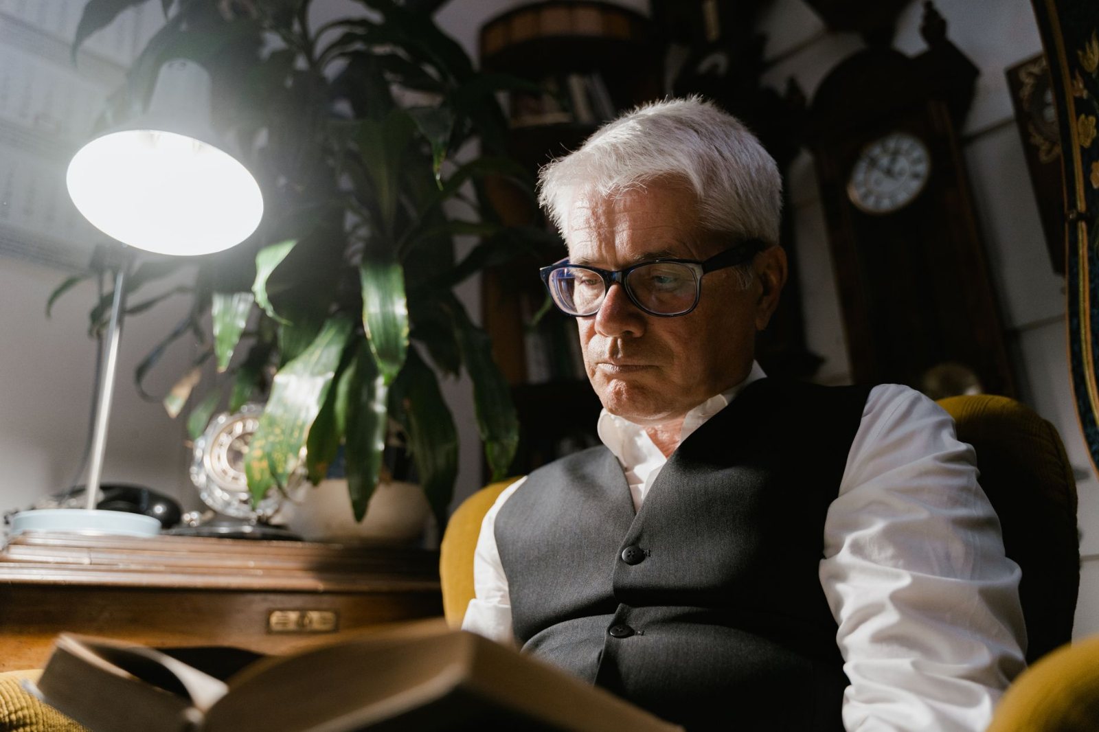An older adult settled comfortably in an armchair in their own front room, lamp switched on against the fading afternoon